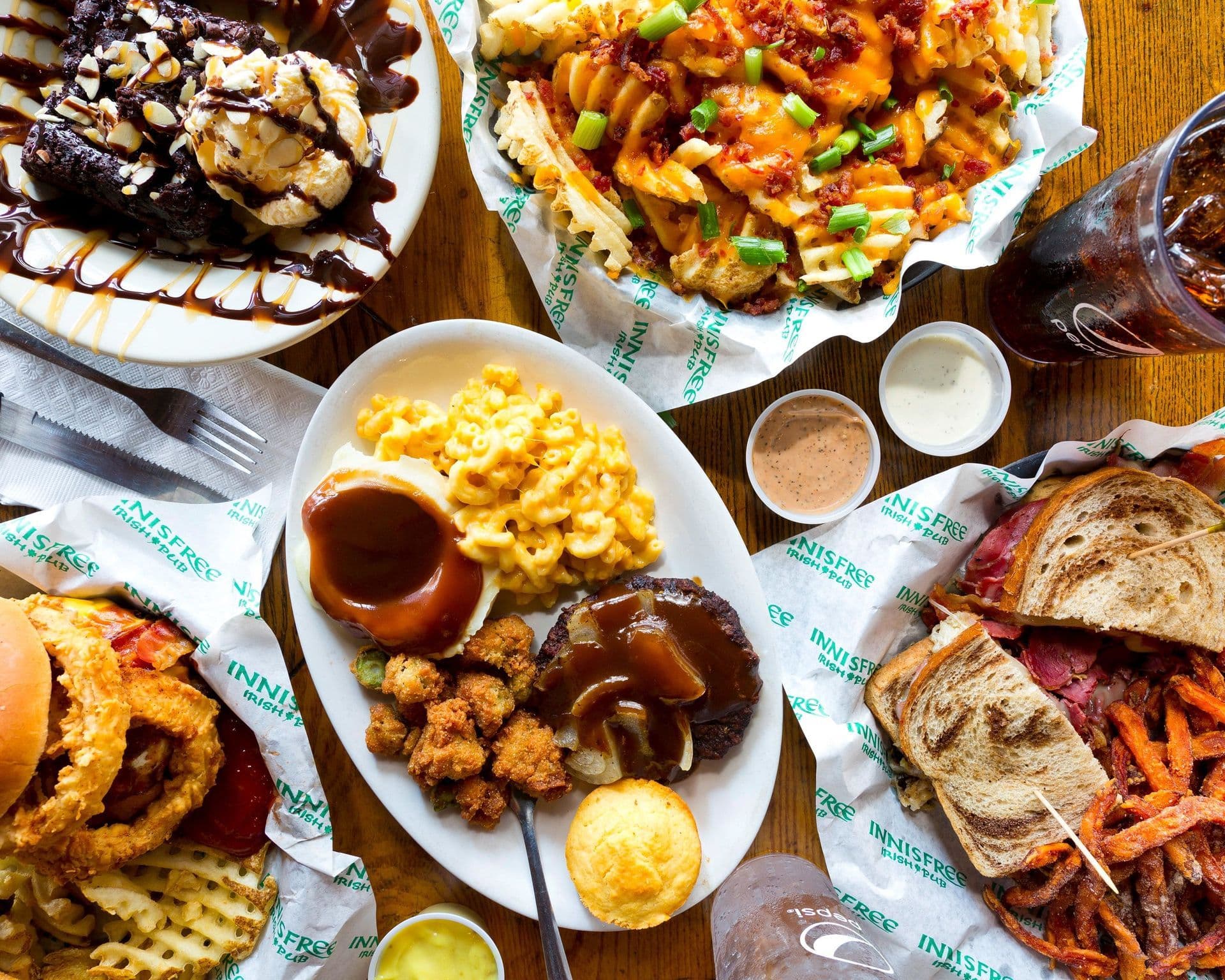 Overhead spread of Innisfree favorites: loaded waffle fries, a meat-and-three plate, a Reuben with sweet potato fries, an onion-ring burger and a brownie sundae